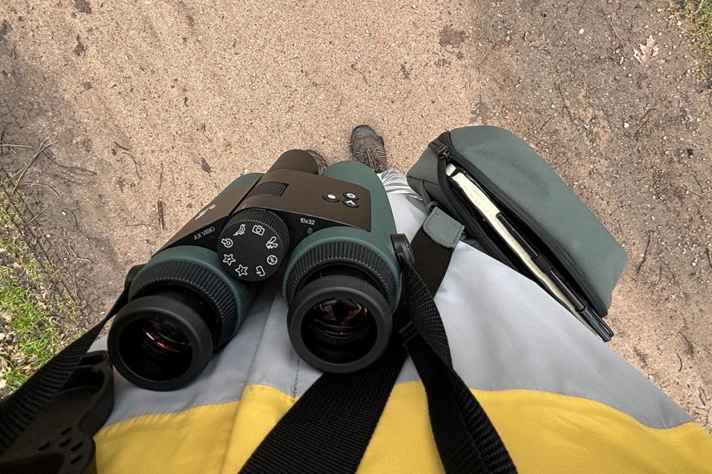 Ein Blick aus Ego-Sicht, ein Mann mit gelber Outdoorjacke sieht an sich herab, vor der Brust hängt ein Fernglas.