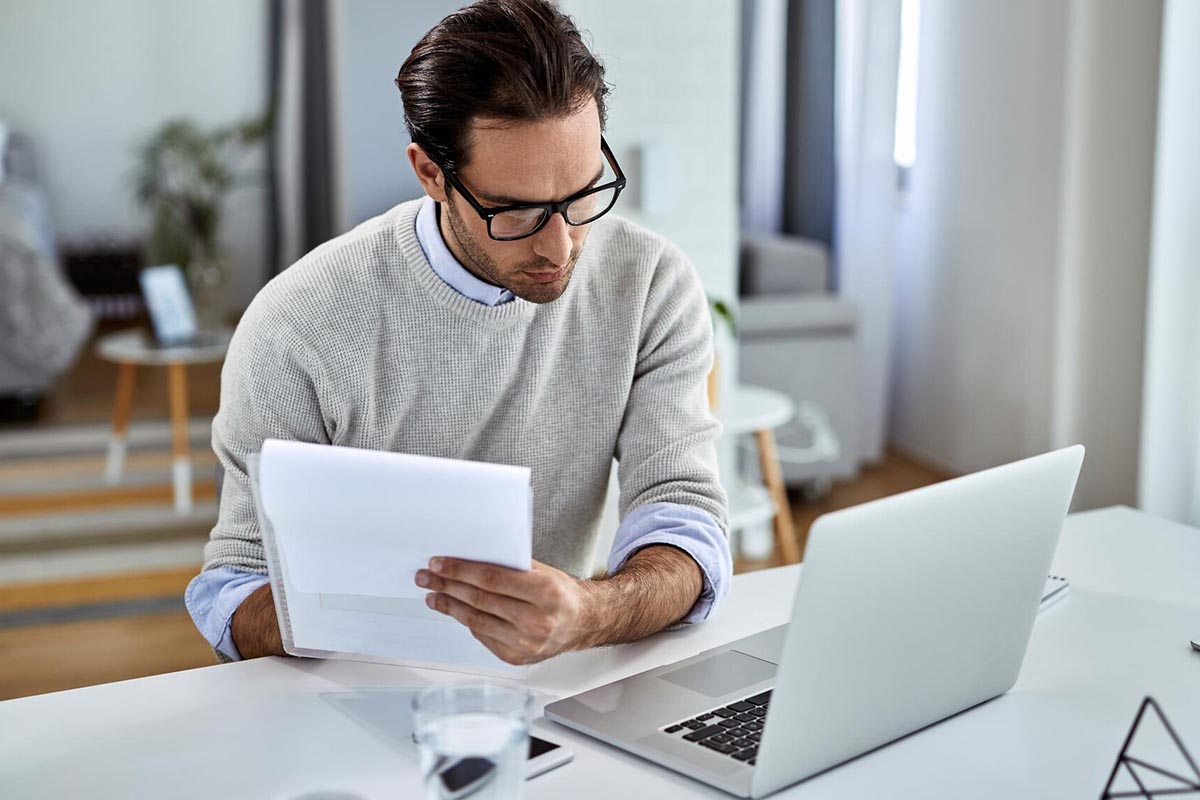 Person an einem Schreibtisch vor einem Laptop mit Zetteln in der Hand.