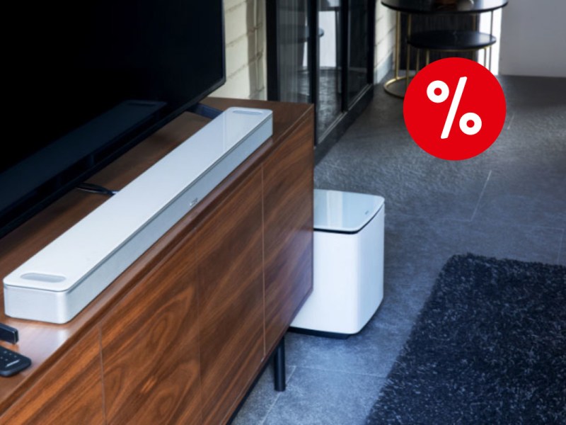 Weiße Soundbar schräg auf dunklem Holzsideboard auf dem schwarzer Fernseher steht auf grauem Boden mit schwarzem Teppich und rotem Prozentbutton oben rechts