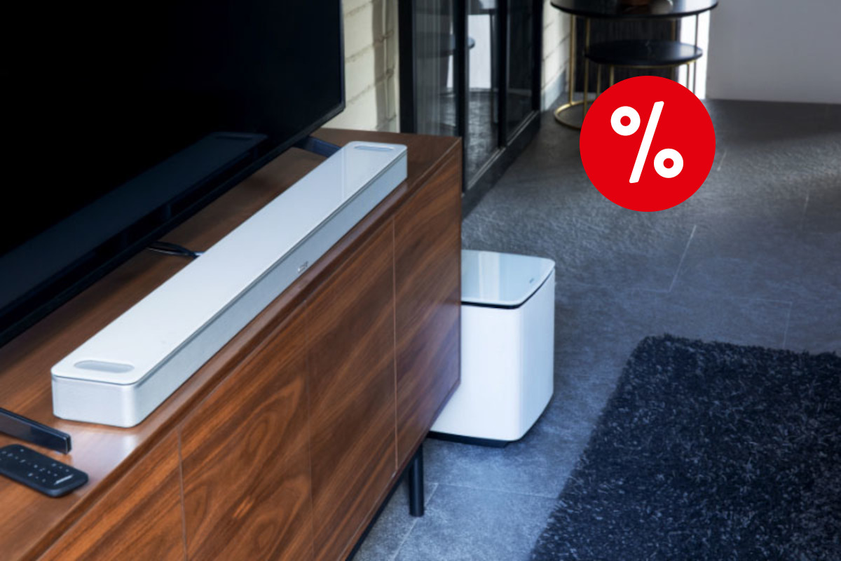 Weiße Soundbar schräg auf dunklem Holzsideboard auf dem schwarzer Fernseher steht auf grauem Boden mit schwarzem Teppich und rotem Prozentbutton oben rechts