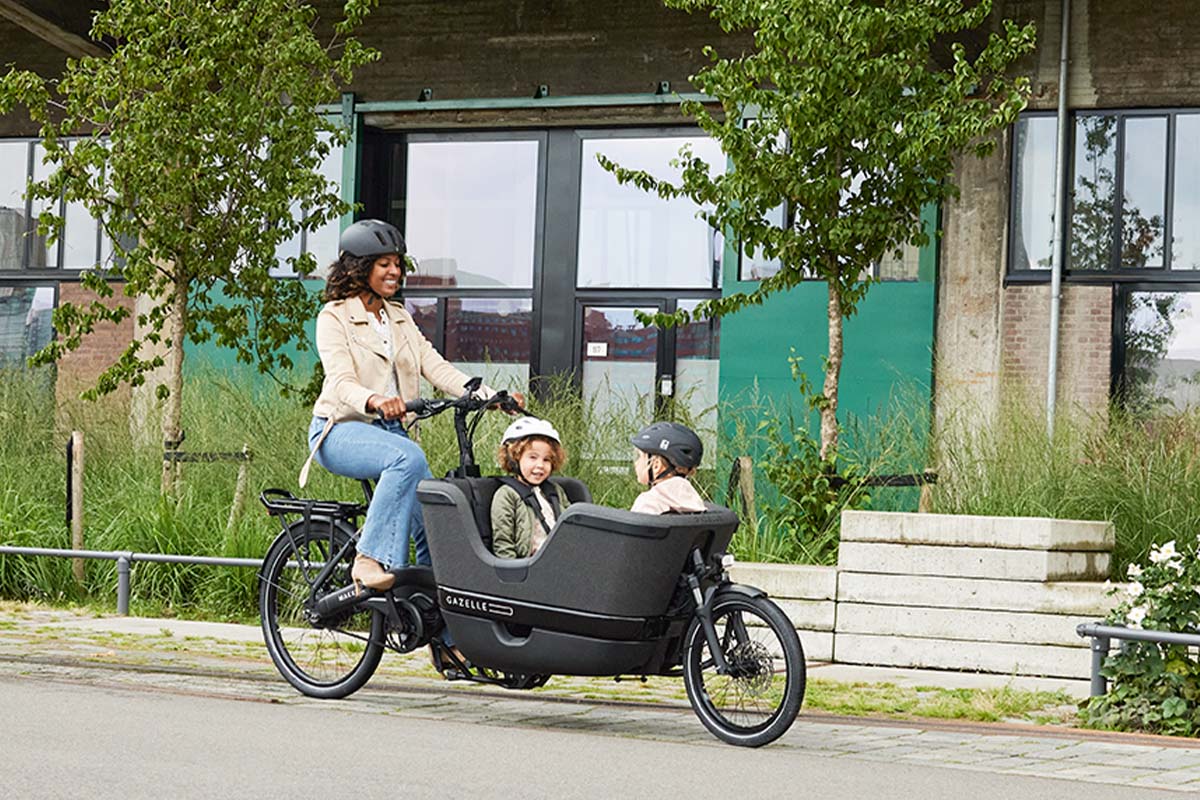 Frau fährt mit einem Lastenrad, in dem zwei Kinder sitzen, durch eine Wohngegend