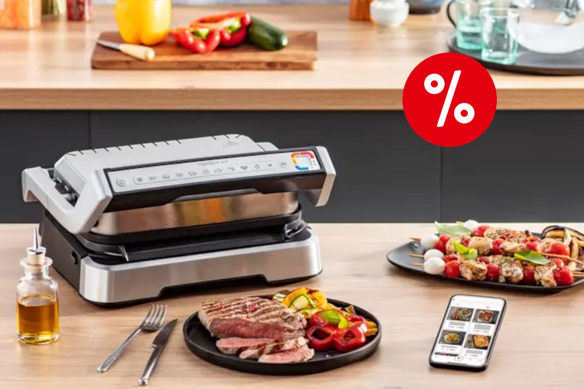 Silberner Tefal Optigrill zugeklappt links auf heller Küchenarbeitsfläche, davor schwarzer Teller mit gegartem Fleisch und gemüse, Besteck, Ölfläschchen, Smartphone mit Rezeoteapp oben rechts ein roter Prozentbutton