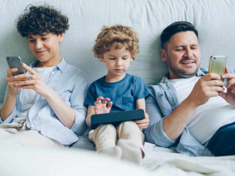 Dunkelhaariger Mann lächelnd rechts liegt in weißem Bett mit hellem Smartphone in der Hand, daneben ein kleiner Junge mit hellen Locken mit Tablet und daneben ein älterer Junge mit dunklen Locken mit Smartphone, lächelnd