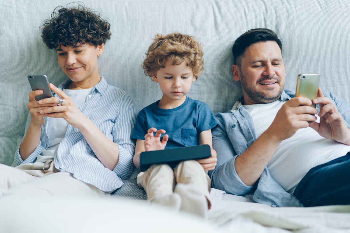 Dunkelhaariger Mann lächelnd rechts liegt in weißem Bett mit hellem Smartphone in der Hand, daneben ein kleiner Junge mit hellen Locken mit Tablet und daneben ein älterer Junge mit dunklen Locken mit Smartphone, lächelnd