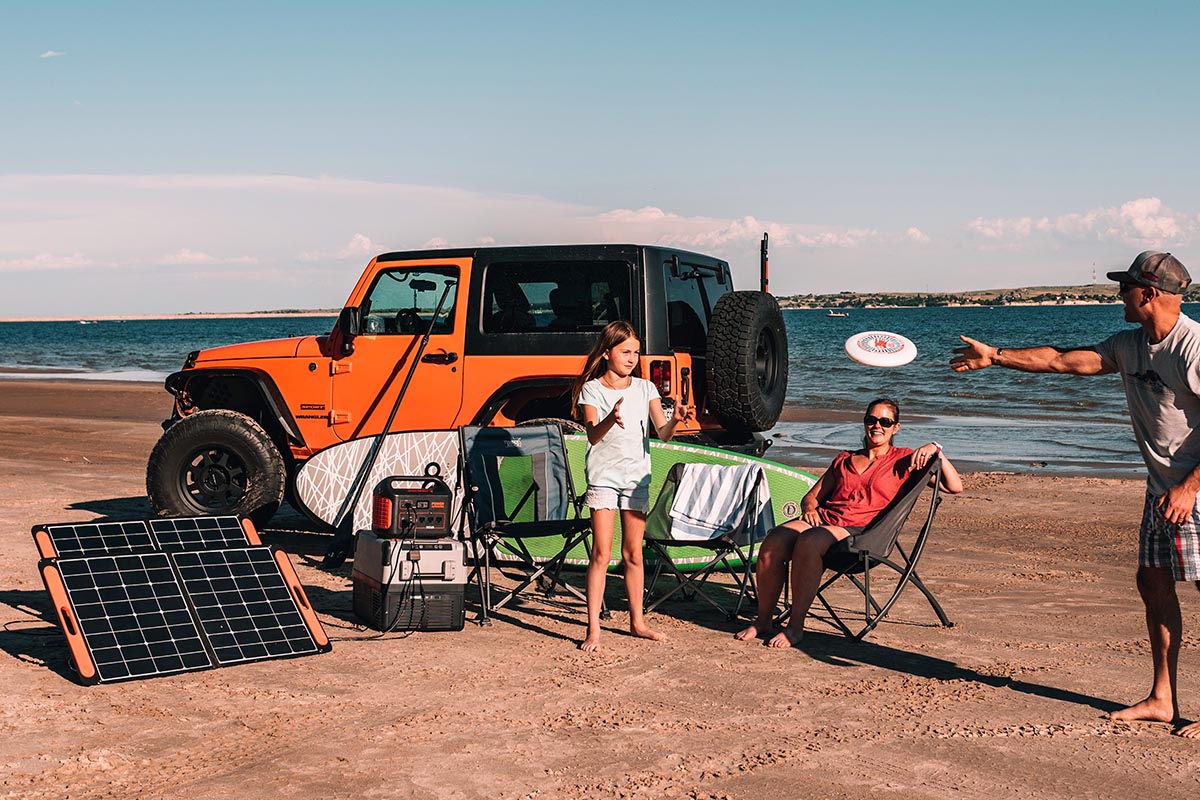 Familie spielt Frisbee am Strand vor orangenem Geländewagen, aufgestellten Solarpanels, Powerstation und Surfbrett