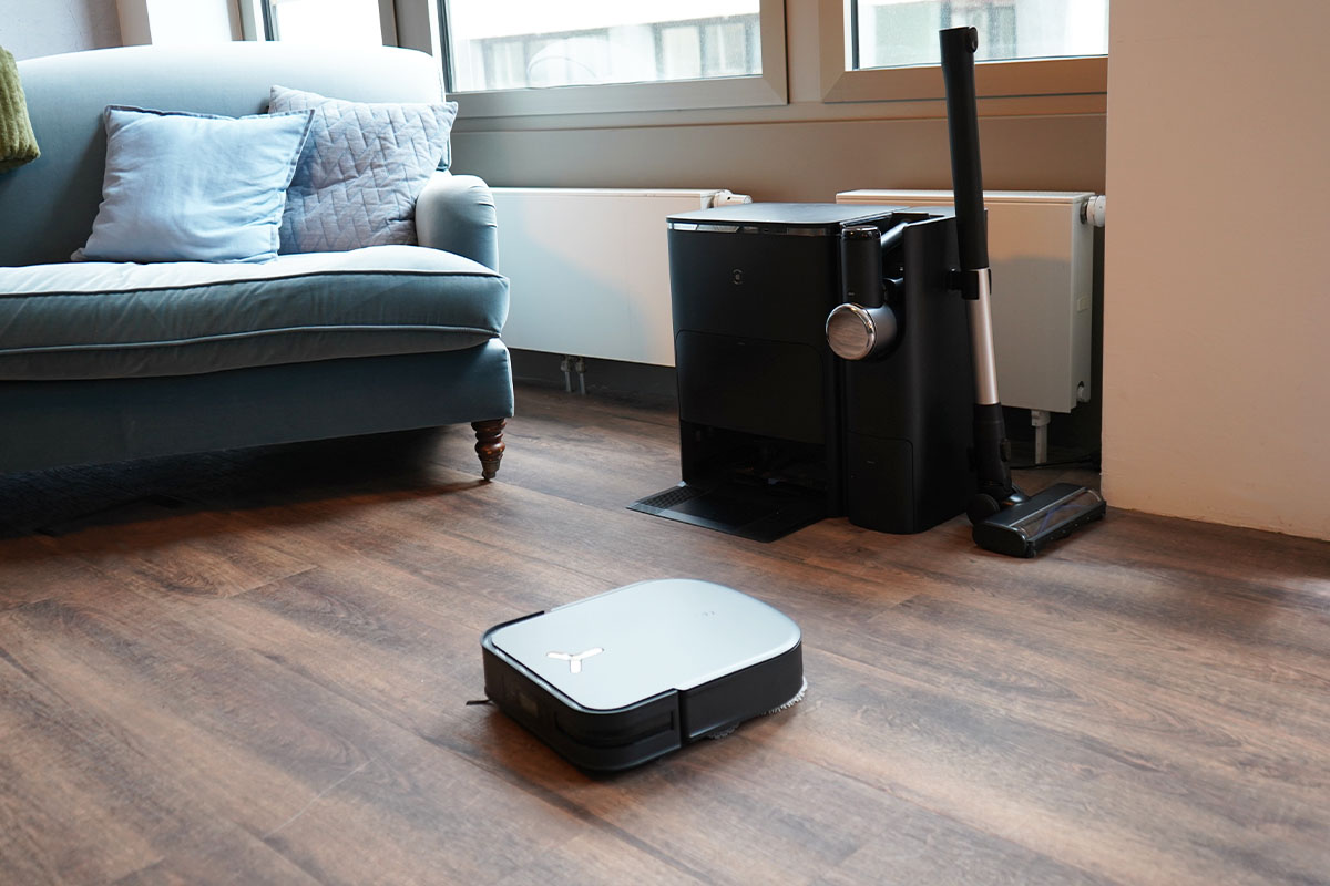 Der Ecovacs Deebot X2 Saugwischroboter in Wohnzimmer-Umgebung. Im Hintergrund steht die Combo-Station mit Handstaubsauger und Akkusauger-Aufsatz.