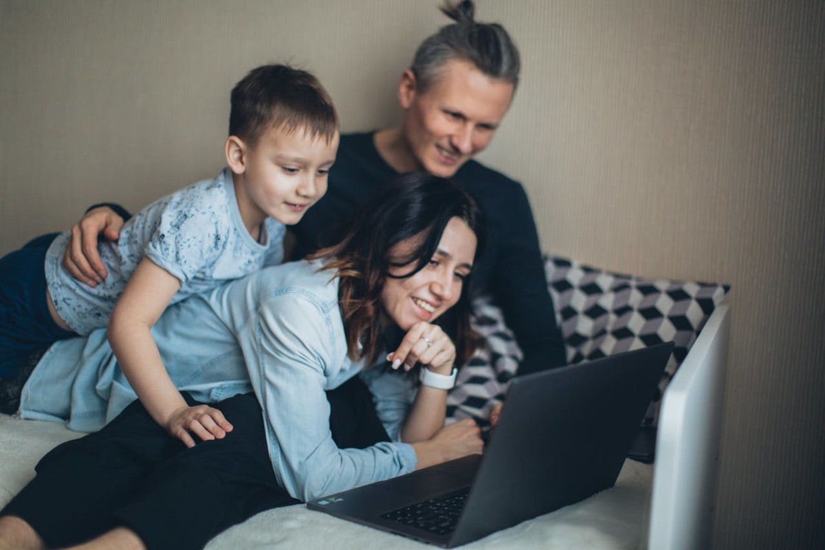 Mann, Frau und Kind, alle dunkelhaarig liegend auf aufgeklapptem schwarzen Laptop guckend, lächelnd vor beiger Wand