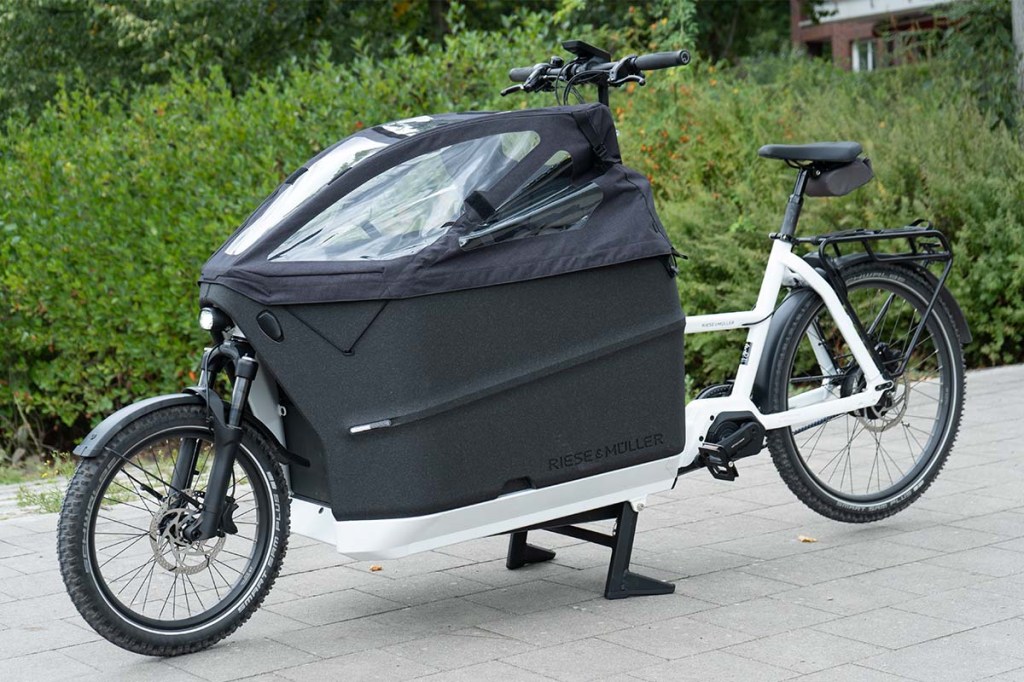 Cargo-E-Bike mit Regenhaube über der Transportwanne, vor einer Hecke stehend