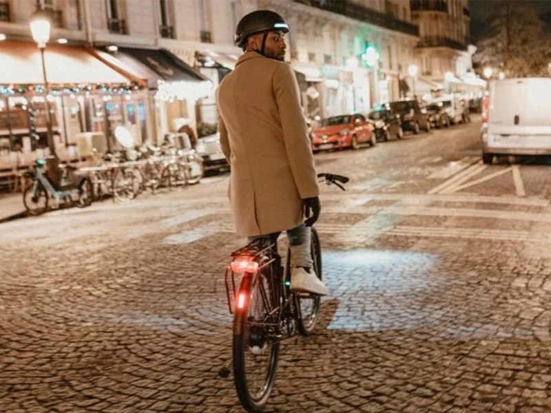 Mann fährt auf seinem Fahrrad durch eine nächtliche Stadt, man sieht ihn von hinten, er trägt einen Helm