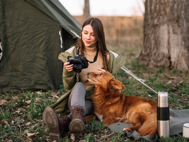 Person mit Hund sitzt auf Gras vor einem Zelt.