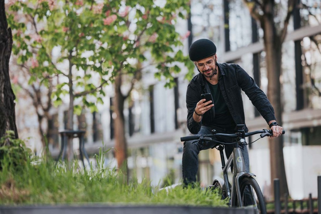 Man sitzt auf seinem Fahrrad, ein Bein auf eine Mauer gelehnt und schaut auf sein Smartphone