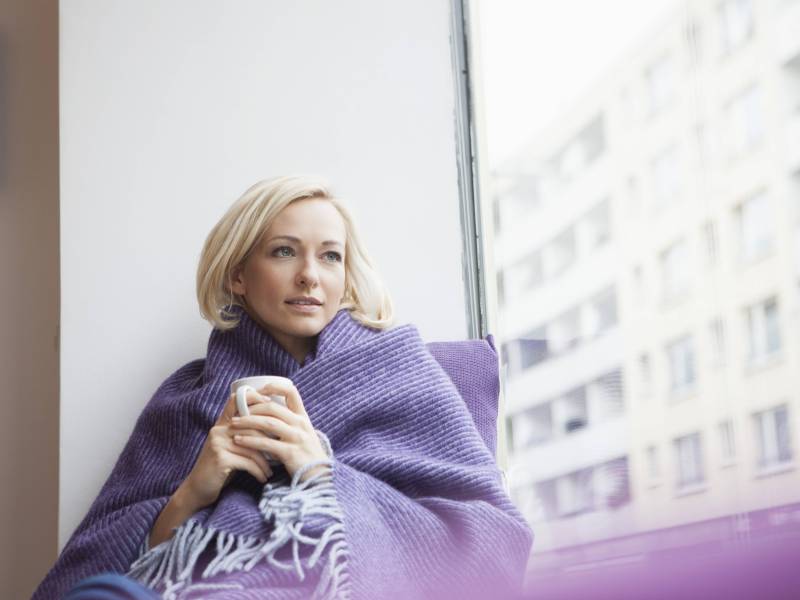 Blonde Frau mit Kurzhaarfrisur sitzt in eine Decke eingehüllt am Fenster und hält eine Tasse in den Händen, während sie gedankenversunken aus dem Fenster schaut.