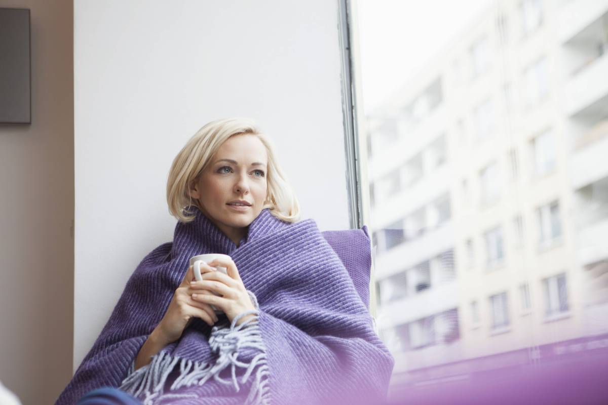 Blonde Frau mit Kurzhaarfrisur sitzt in eine Decke eingehüllt am Fenster und hält eine Tasse in den Händen, während sie gedankenversunken aus dem Fenster schaut.