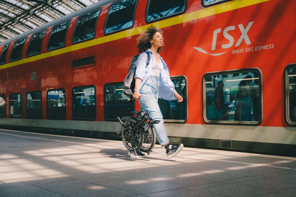 Frau geht mit einem Faltrad einen Bahnsteig entlang, Bahn im Hintergrund