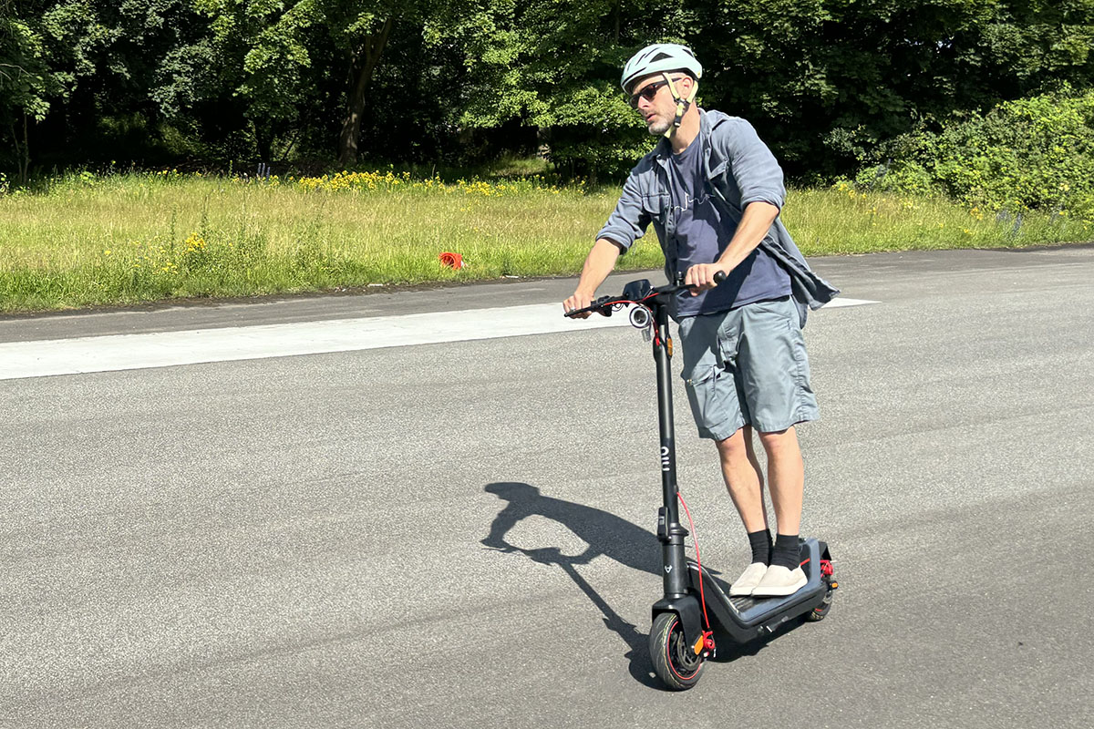 Mann fährt auf einem E-Scooter. Man sieht ihn kurvenfahrend an einem Waldstück vorbeifahrend.