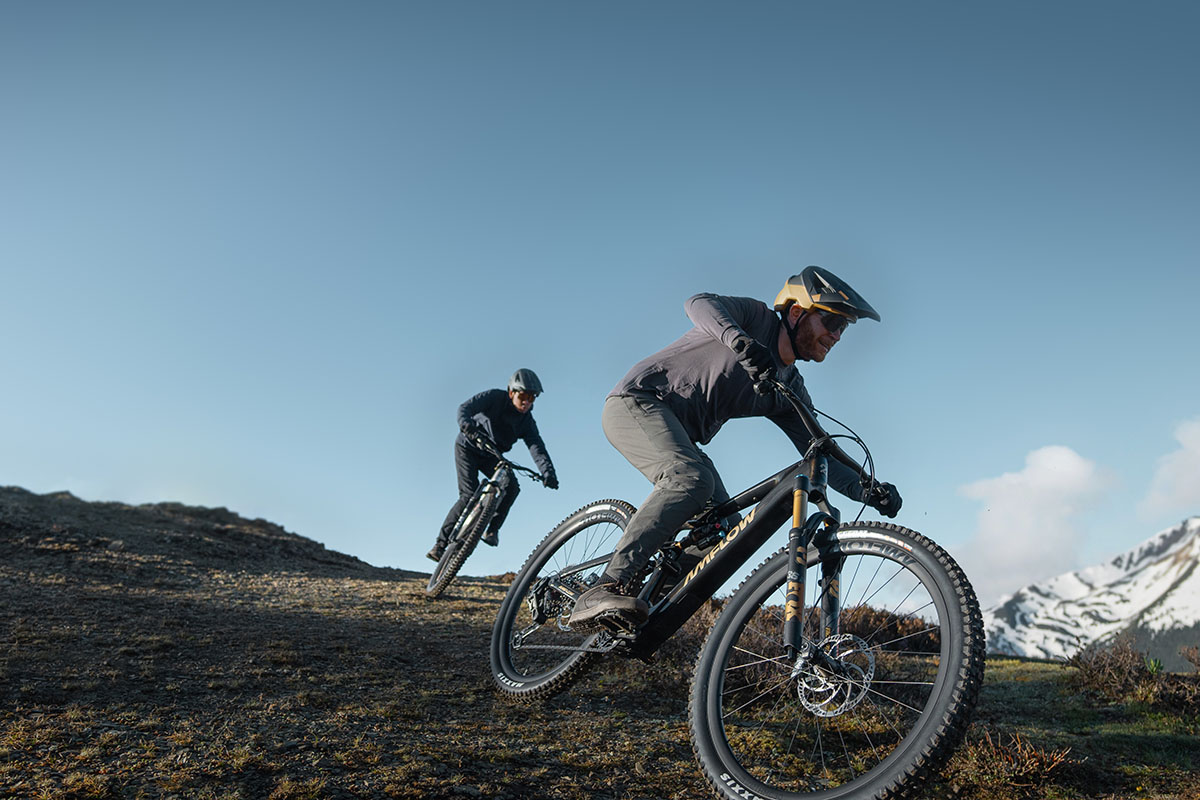 Zwei Männer fahren auf E-Mountainbikes einen Hang hinab. Dabei tragen beide je einen Sturzhelm.