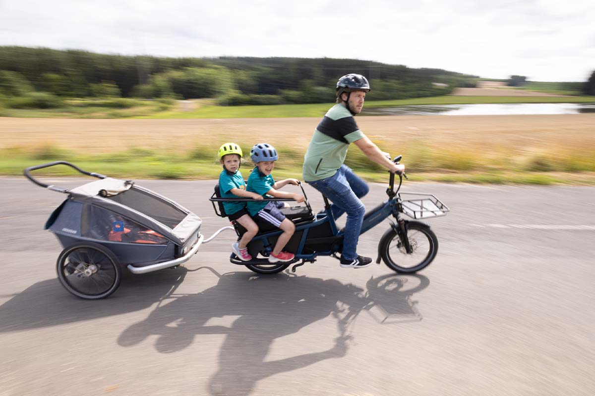 Cargo-E-Bike mit Vater und zwei Kindern, dahinter ein Anhänger, fahren auf einer Landstraße