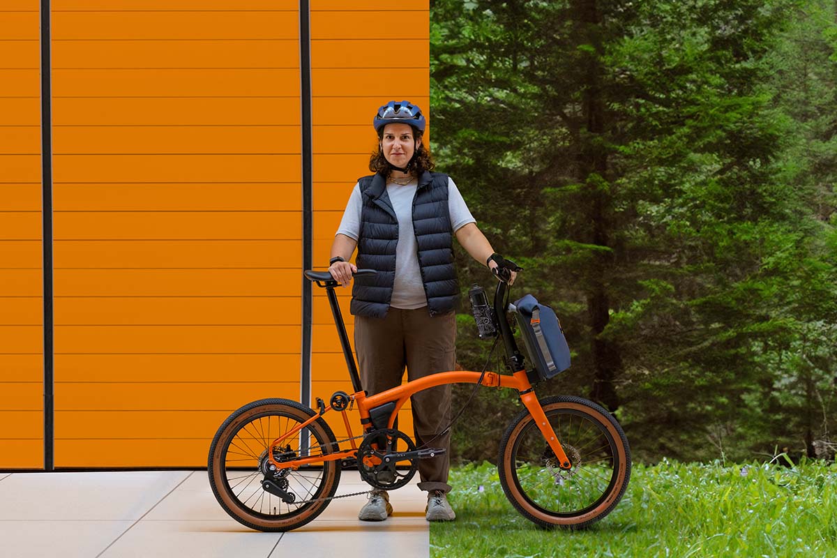 Frau steht hinter ihrem orangenen Faltrad, eine orangene Wand zu einem Teil im HIntergrund