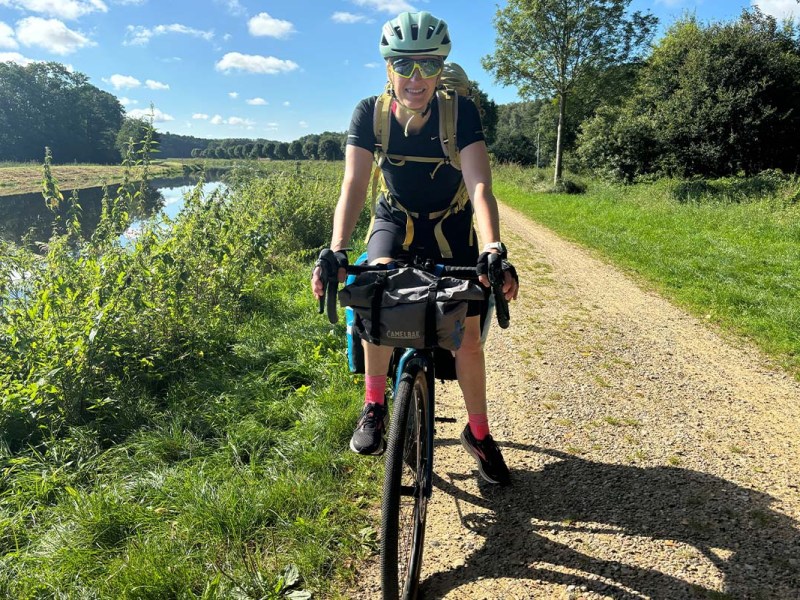 Frau von vorne auf einem bepackten Gravelbike, Natur und ein Kanal im HIntergrund