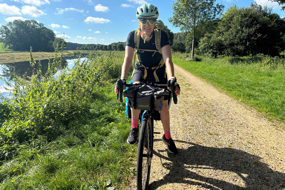 Frau von vorne auf einem bepackten Gravelbike, Natur und ein Kanal im HIntergrund