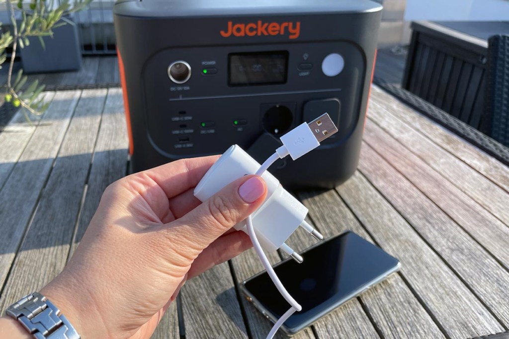 Eine Hand hält ein USB-Kabel und ein Netzteil vor die Jackery Explorer 1000 v2 und ein Handy auf einem Tisch.