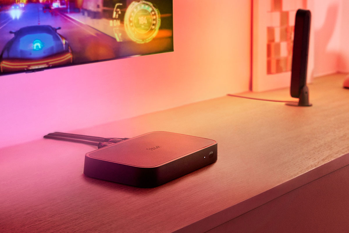 Streaming-Box Philips Hue Play HDMI Sync Box 8K liegt auf einem Sideboard unter einem Flachbild-TV.