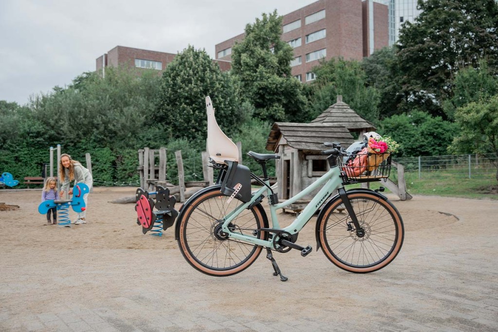 türkisfarbenes E-Bike mit Kindersitz und Frontgepäckträger vor einem Spielplatz stehend, Mutter mit Kind im Hintergrund