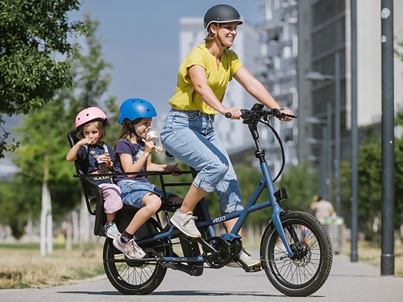 Frau fährtb auf klappbaren E-Cargo-Bike Vello Sub auf einer Straße ein einer Stadt. Auf dem Gepäckträger befinden sich zwei Kindersitze, in denen Kinder sitzen.