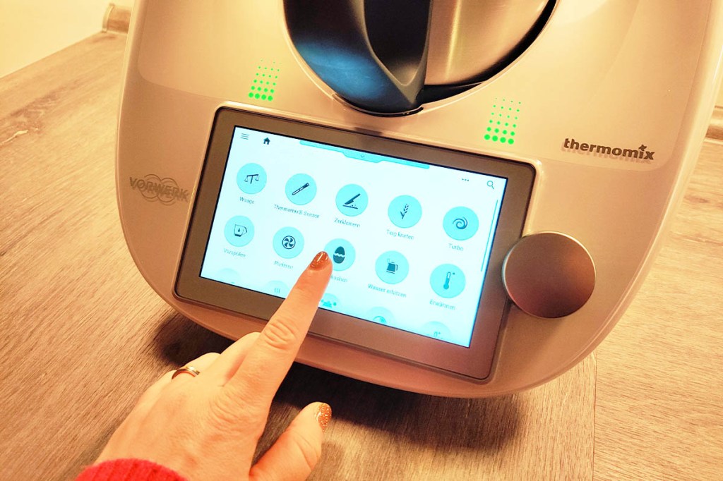 Eine Hand bedient das Display des Thermomix TM6, auf dem Funktionen zu sehen sind.